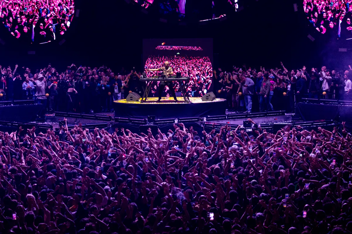 Vista general del Movistar Arena con el público de brazos en alto frente a la cabina de Fatboy Slim, iluminado en violeta