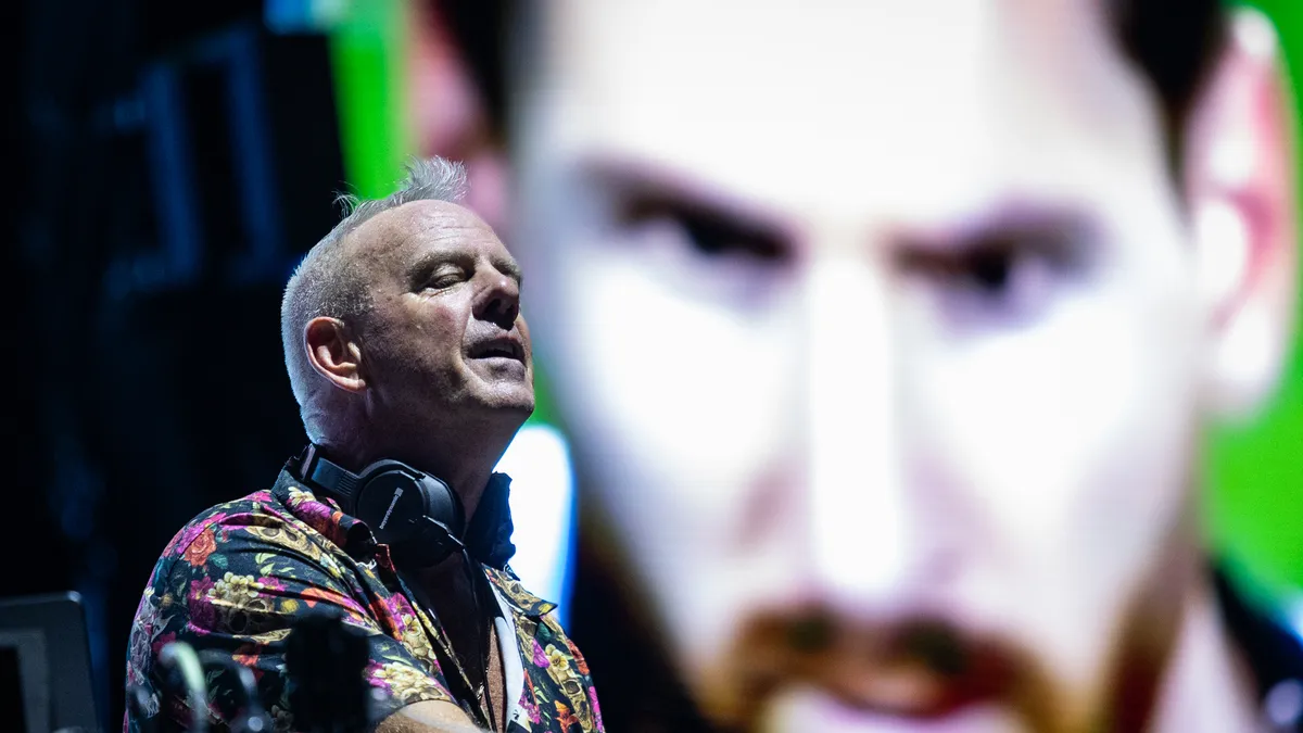 Norman Cook con camisa floreada y auriculares al cuello, de perfil, frente a una pantalla gigante con el rostro de Lionel Messi